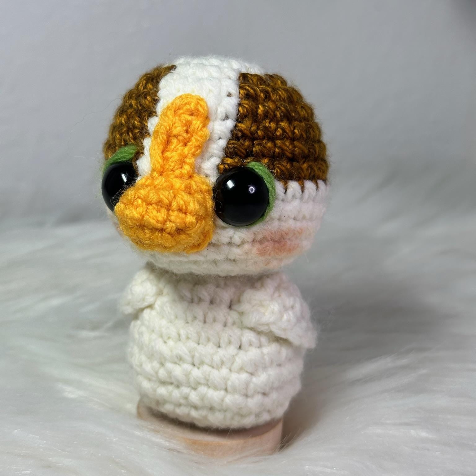 Crocheted animal figurine with a white body, brown and orange head, and black eyes on a white background.
