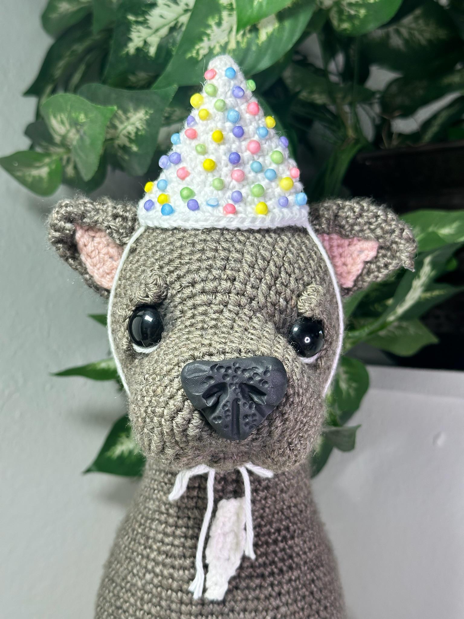 Crocheted animal toy wearing a colorful party hat with a plant in the background