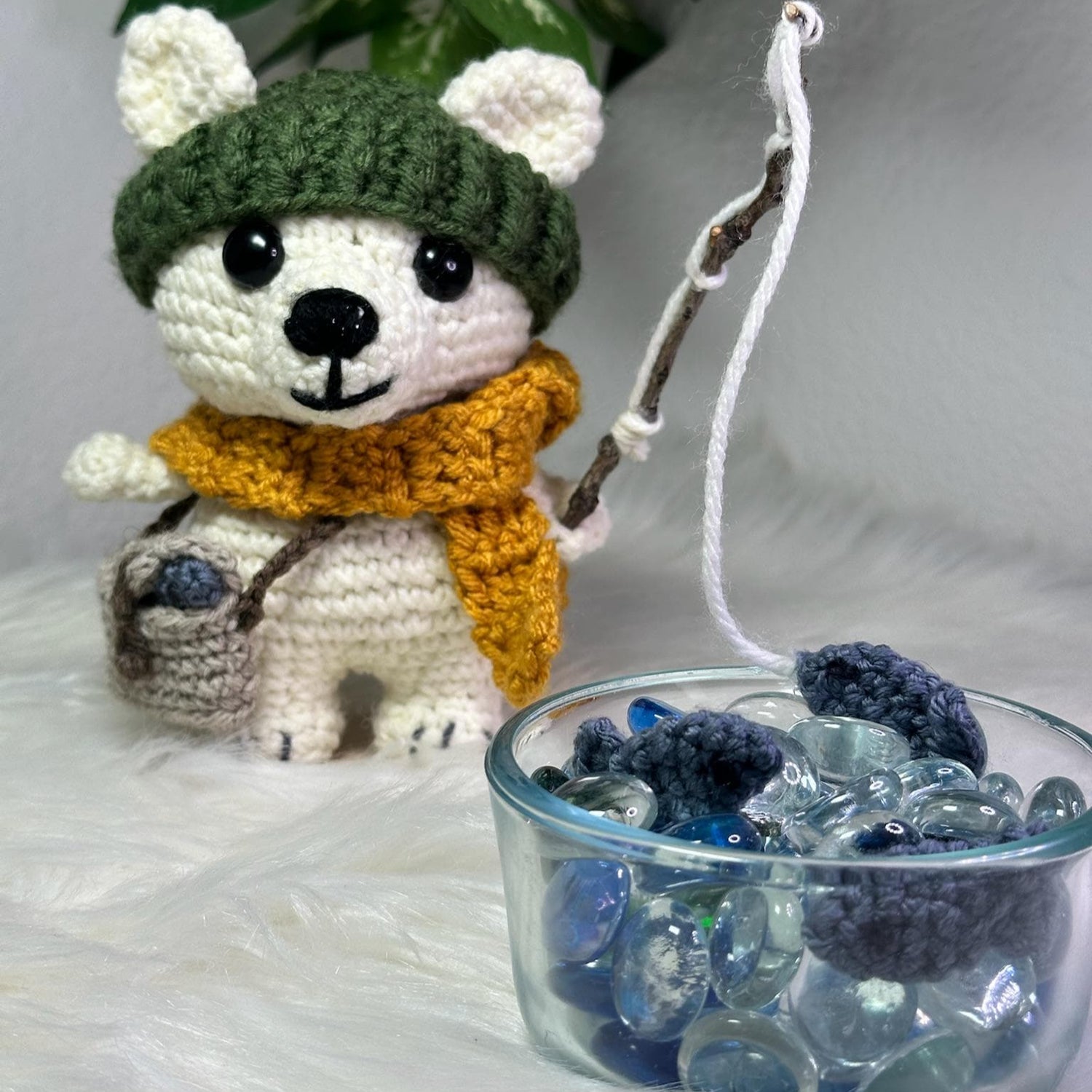 Crocheted bear toy with a green hat and orange scarf next to a glass bowl filled with blueberries on a white surface.