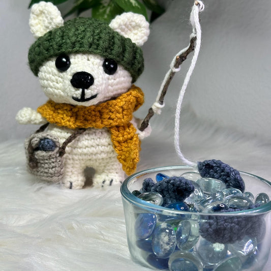 Crocheted bear toy with a green hat and orange scarf next to a glass bowl filled with blueberries on a white surface.